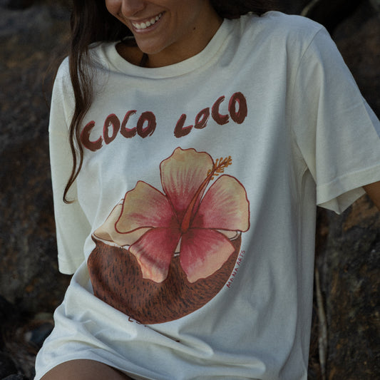 Woman wearing a white t-shirt with a pink flower graphic and 'Coco Loco' text, sitting on rocks.
